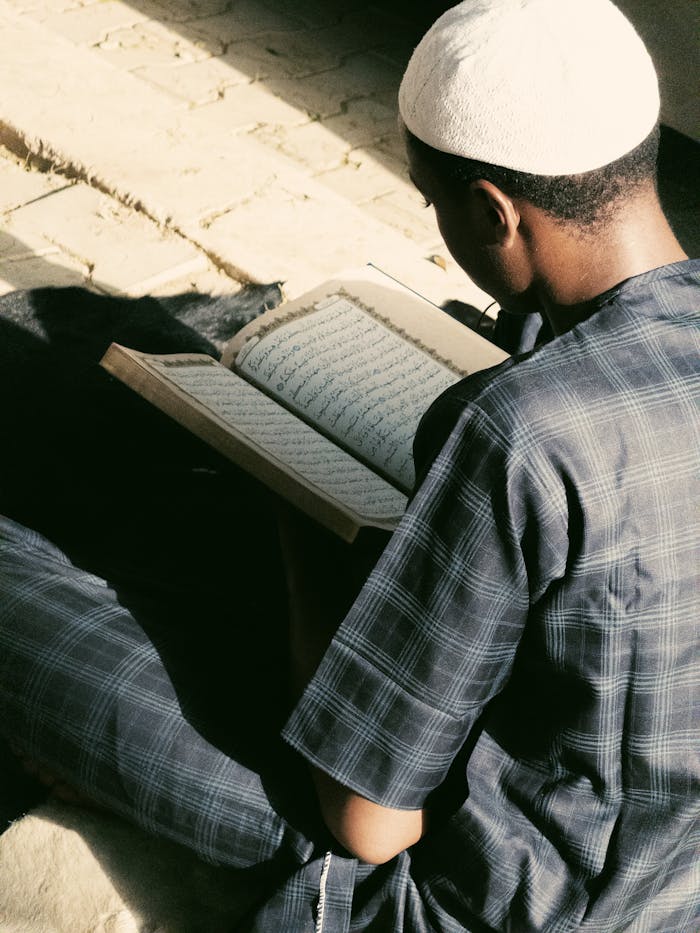 A young man in Kano, Nigeria, engaged in reading the Quran outdoors, capturing a serene moment of reflection.